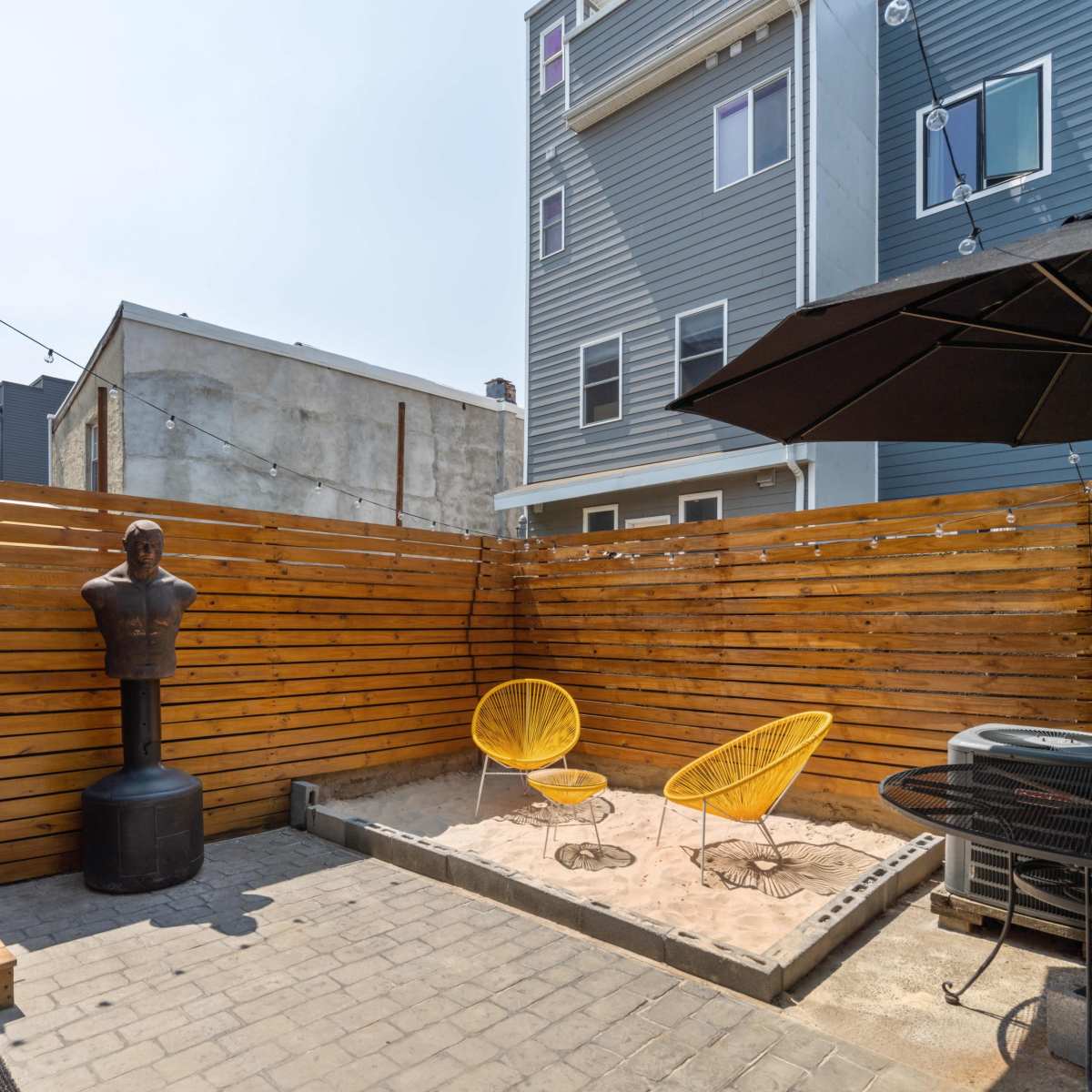A small outdoor patio area featuring a sand pit, two yellow chairs, a black umbrella, and a wooden privacy fence.