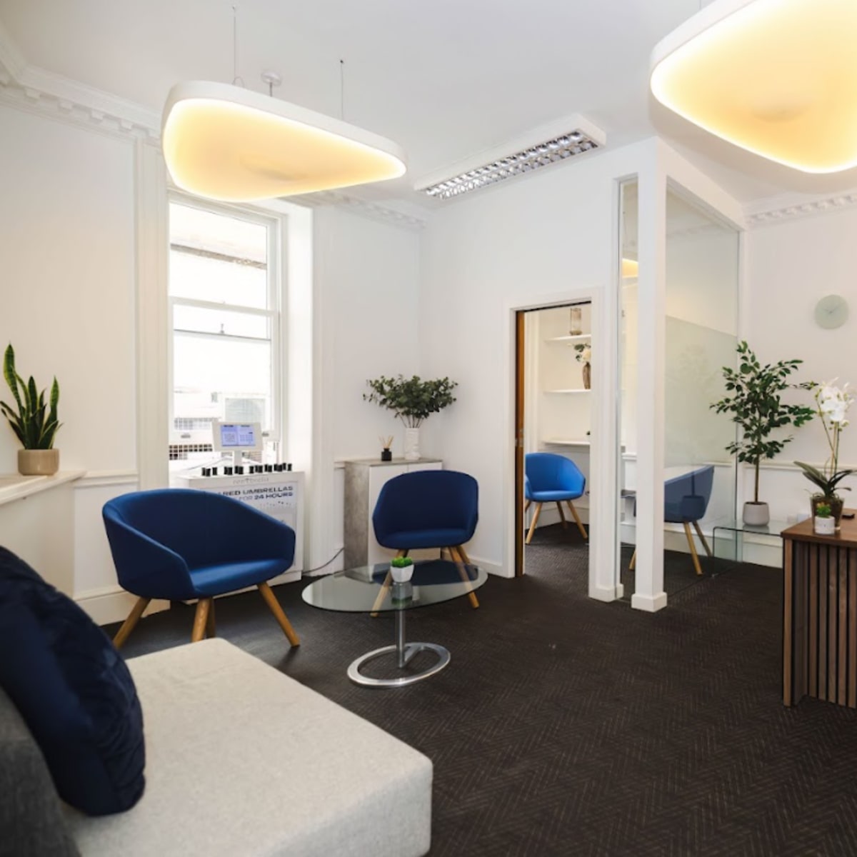A modern office space with two blue chairs, a glass coffee table, a couch, a desk with a computer, and plants in each corner.