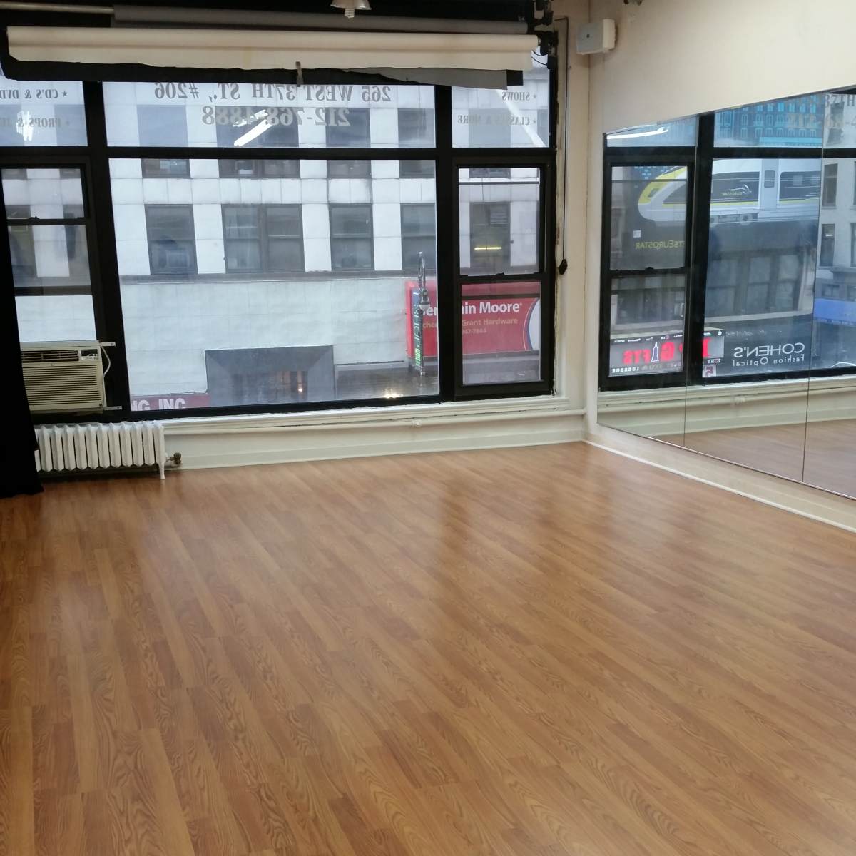 A spacious room with wooden flooring, large windows facing a city street, and a mirrored wall.