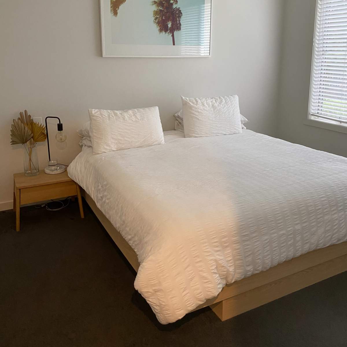 A minimalist bedroom with a large bed, two white pillows, a nightstand, and a framed picture of palm trees on the wall.