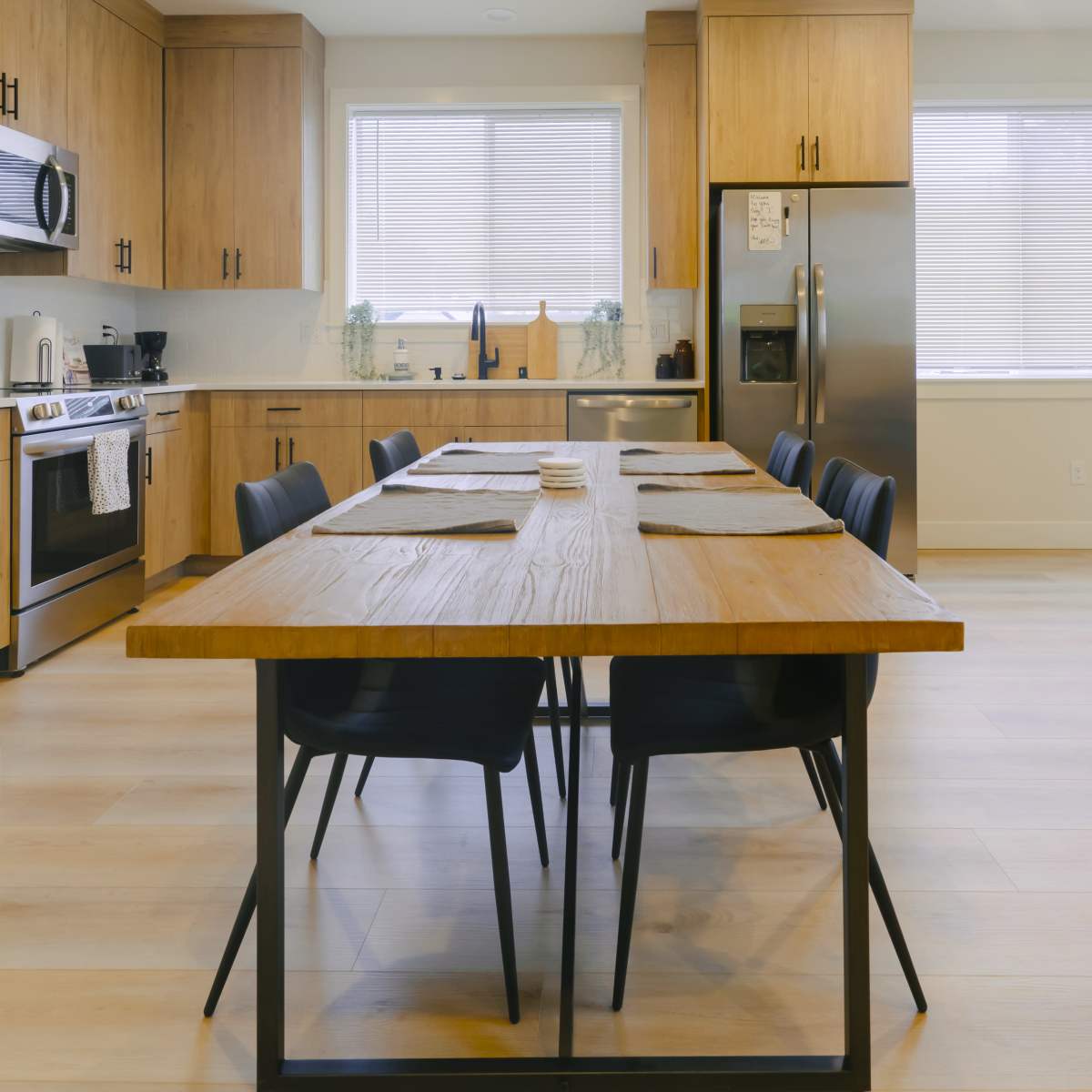 A modern kitchen features wooden cabinetry, stainless steel appliances, and a large dining table with black chairs.
