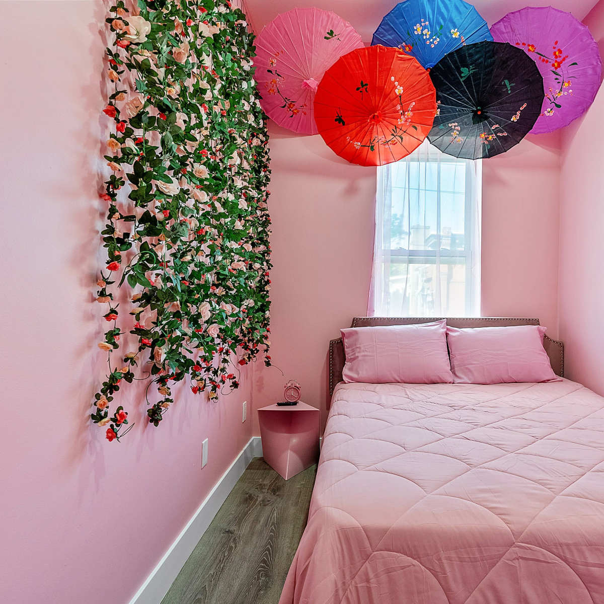 A small bedroom features a pink bedspread, a floral wall decoration, and colorful umbrellas hanging from the ceiling.
