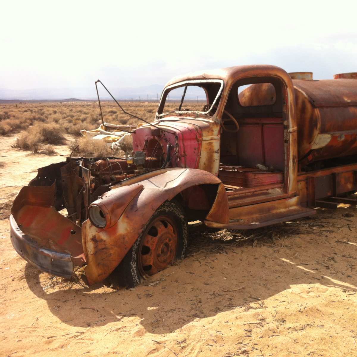 An old, rusted tanker truck is abandoned in a sandy, arid landscape with sparse vegetation.