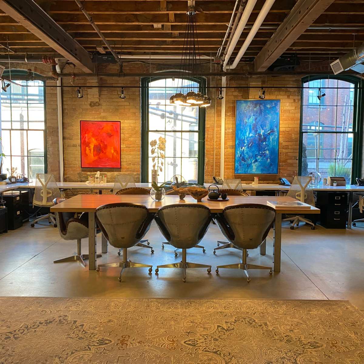 A spacious office with large windows, featuring a long wooden table surrounded by modern chairs and colorful abstract paintings on the walls.