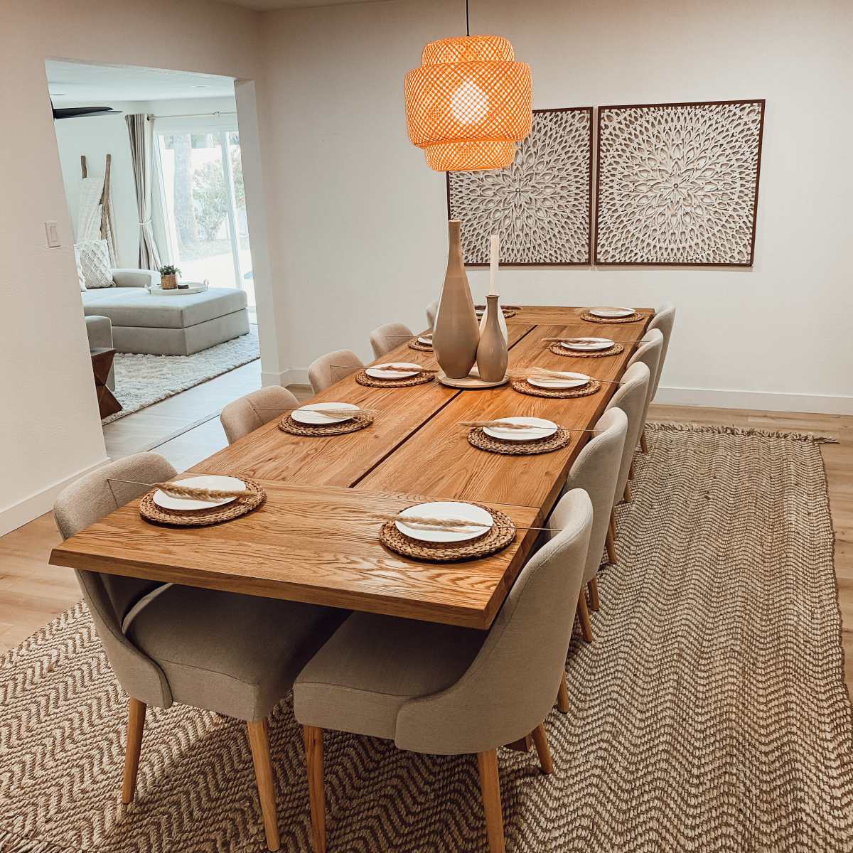 A long wooden dining table is set with plates and utensils, surrounded by upholstered chairs on a woven rug, with a decorative lamp and art pieces in the background.