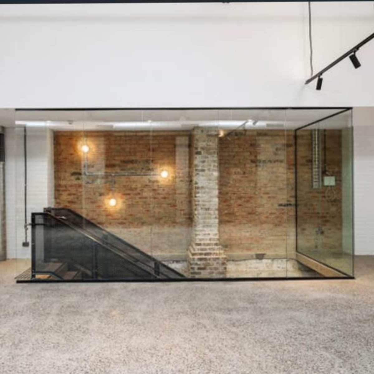 A modern staircase enclosed in glass, featuring a brick wall with exposed light fixtures in a spacious interior.