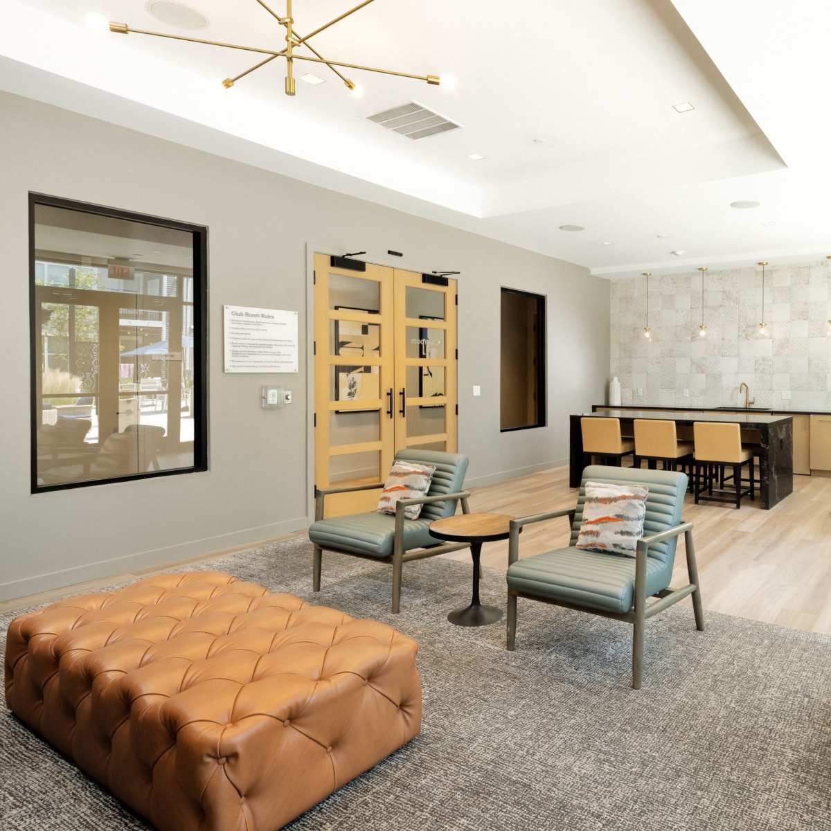 A modern lounge area with a tufted leather ottoman, seating arrangements, a dining table, and a large countertop with bar stools, all set in a well-lit space with large windows.