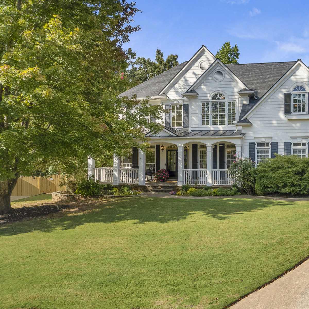 A two-story white house with a covered front porch, surrounded by greenery and a paved driveway.