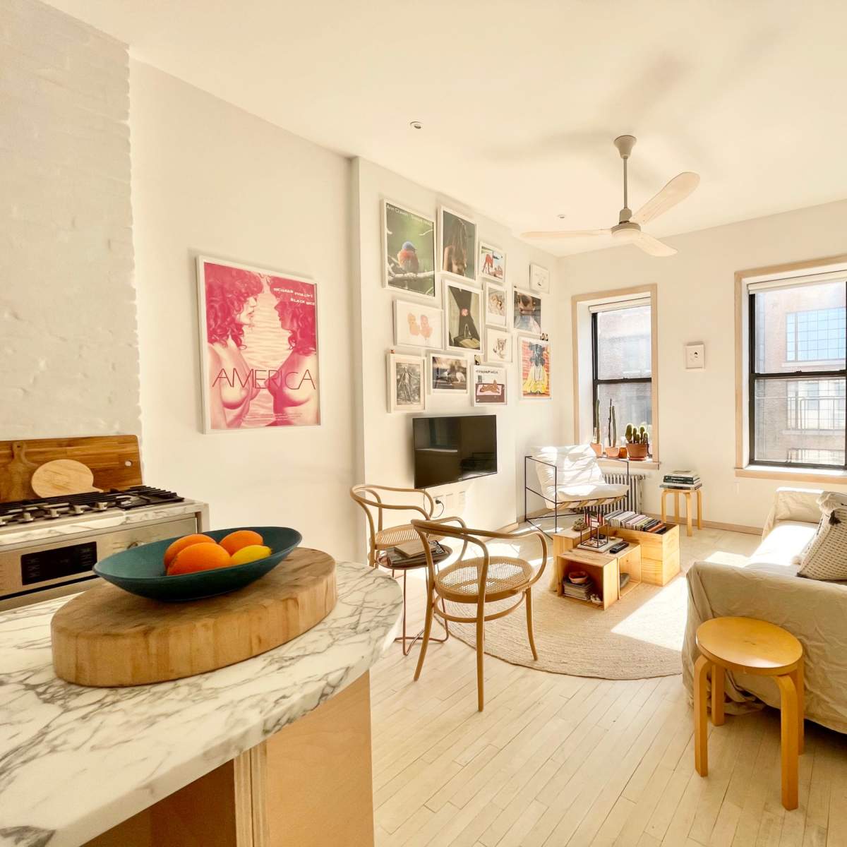 A bright, open-concept living space features a kitchen with a marble countertop, a cozy sitting area with a sofa and decorative plants, and walls adorned with framed art.