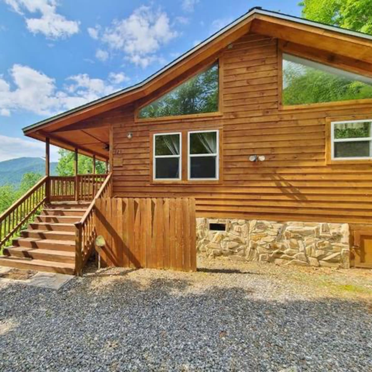 A wooden cabin with a stone foundation and large windows overlooks a mountainous landscape under a clear blue sky.