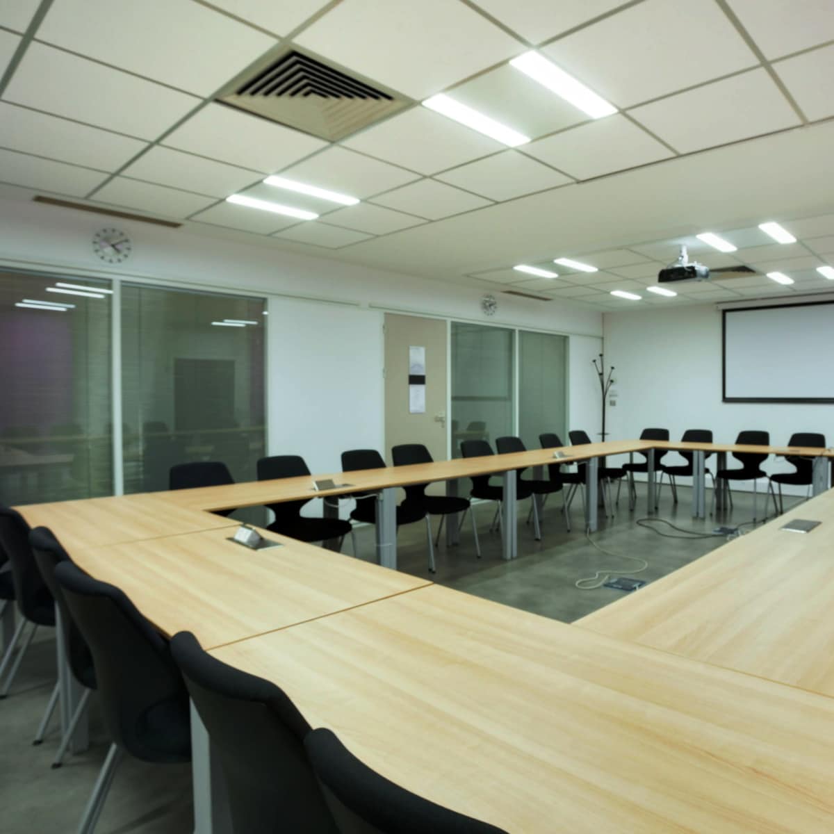 A large conference room features a U-shaped arrangement of wooden tables and black chairs, with a projector screen at one end and glass windows along the walls.