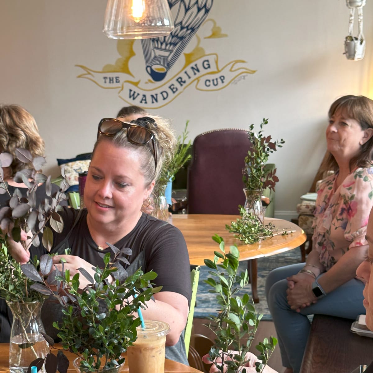 A woman arranges greenery in a glass vase at a table while others observe in a café decorated with a mural.