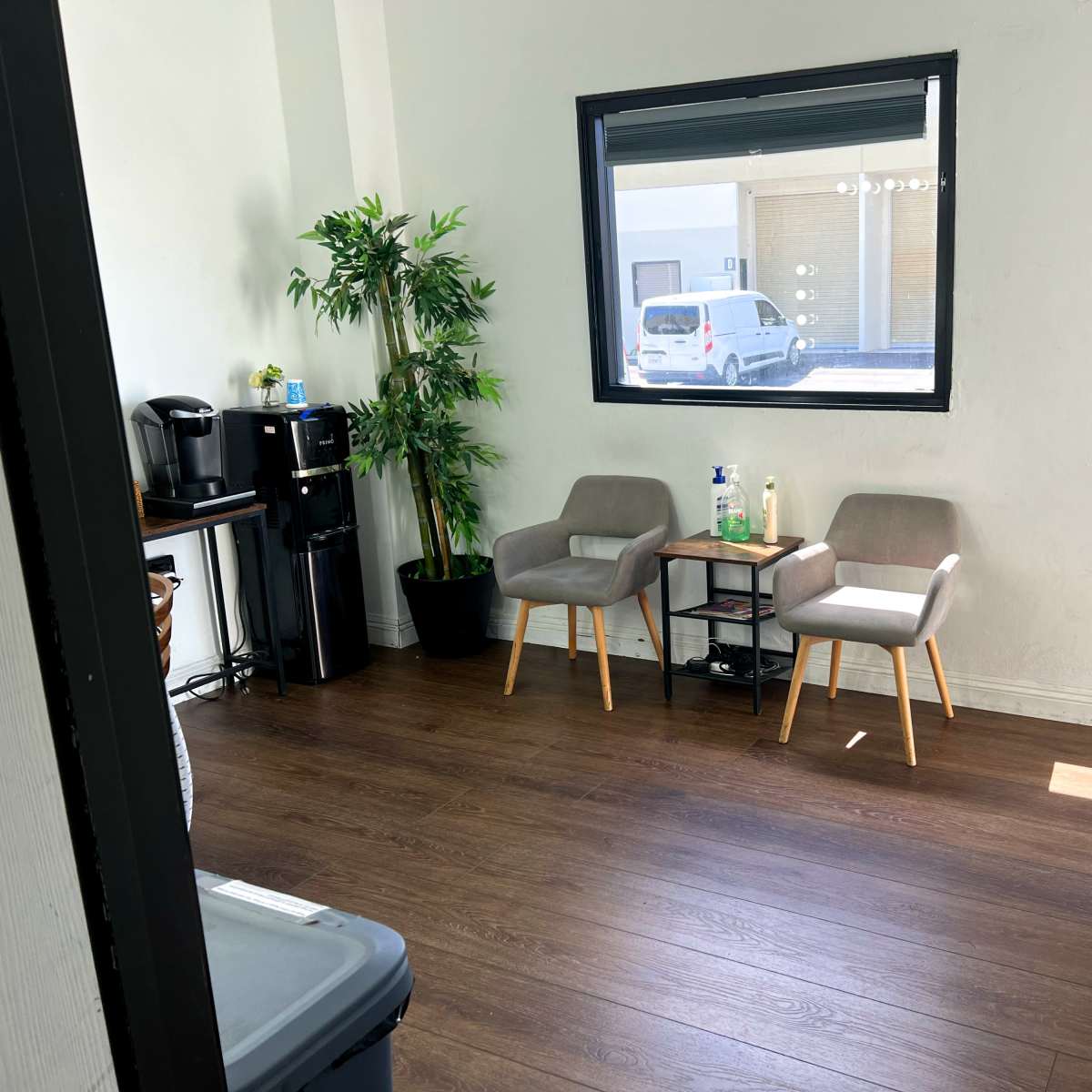 A small room with two gray chairs, a side table, a potted plant, and a coffee station along one wall.
