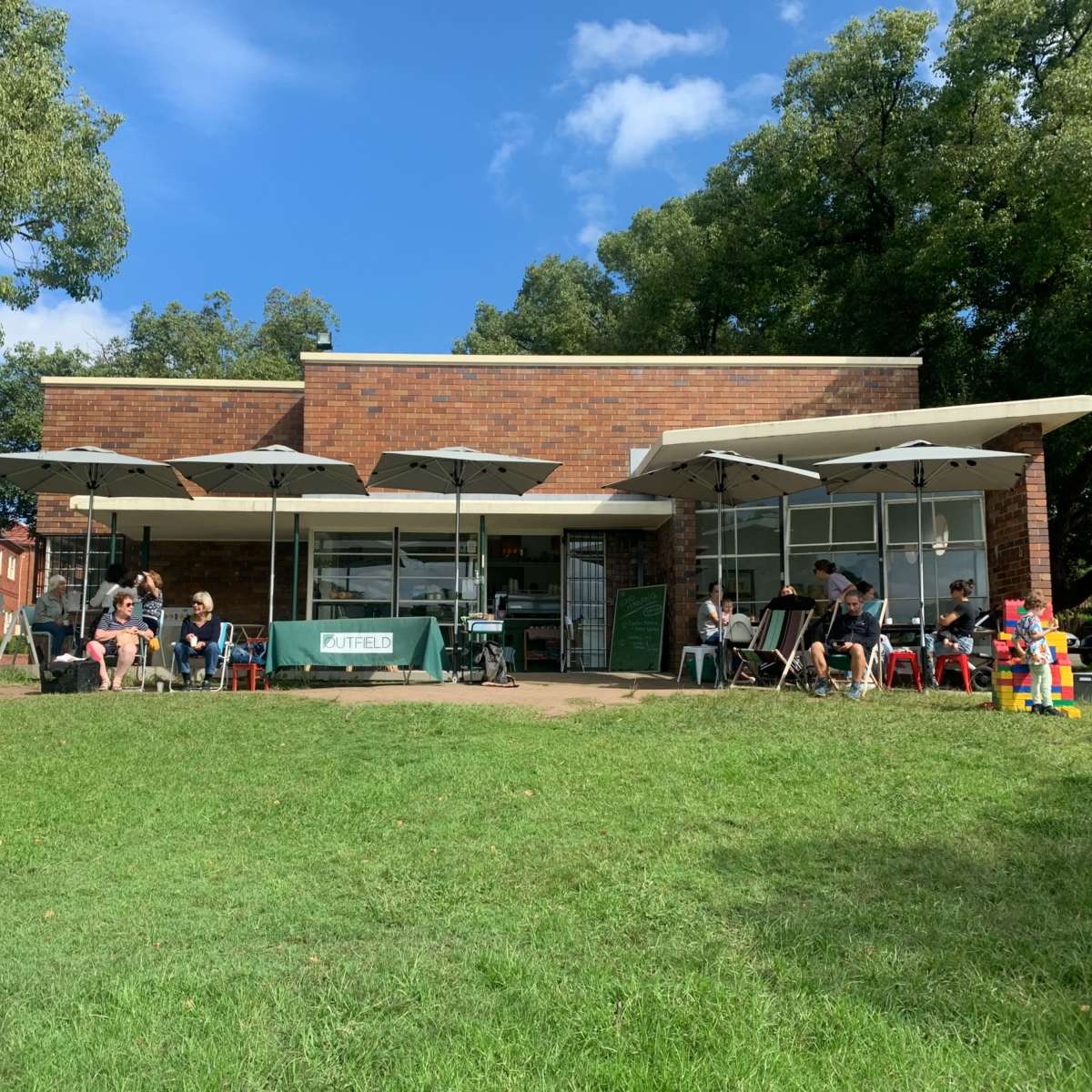 A brick cafe with outdoor seating shaded by umbrellas is situated on a grassy area, surrounded by trees.