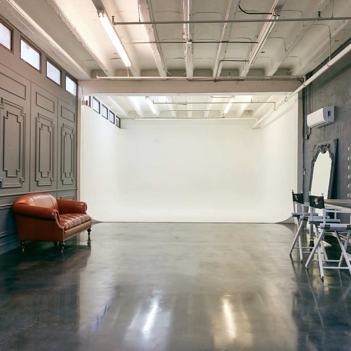A spacious studio with a white backdrop, a leather sofa against a wall, and a mirrored makeup station on wheels.