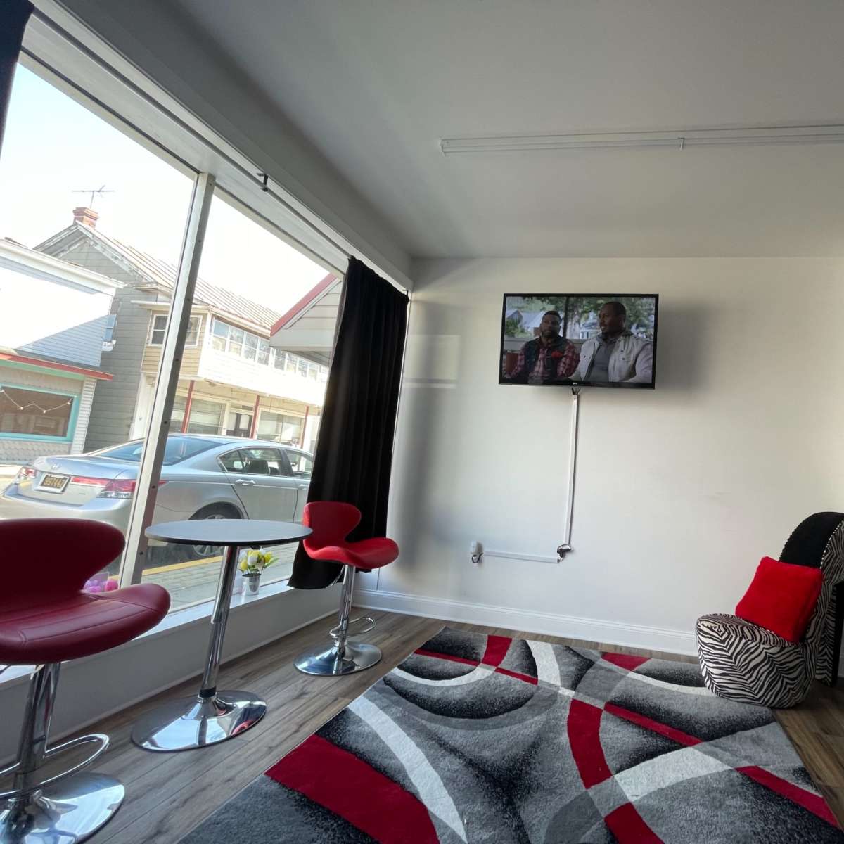 A modern, well-lit room featuring two red barstools, a zebra-striped chair, a large TV mounted on the wall, and a patterned gray rug on the floor.