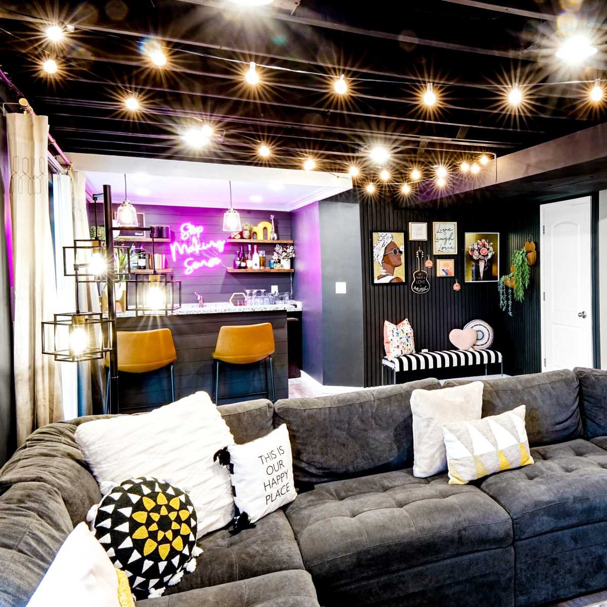 A modern living room features a large sectional sofa, a stylish bar area with purple lighting, and string lights overhead.
