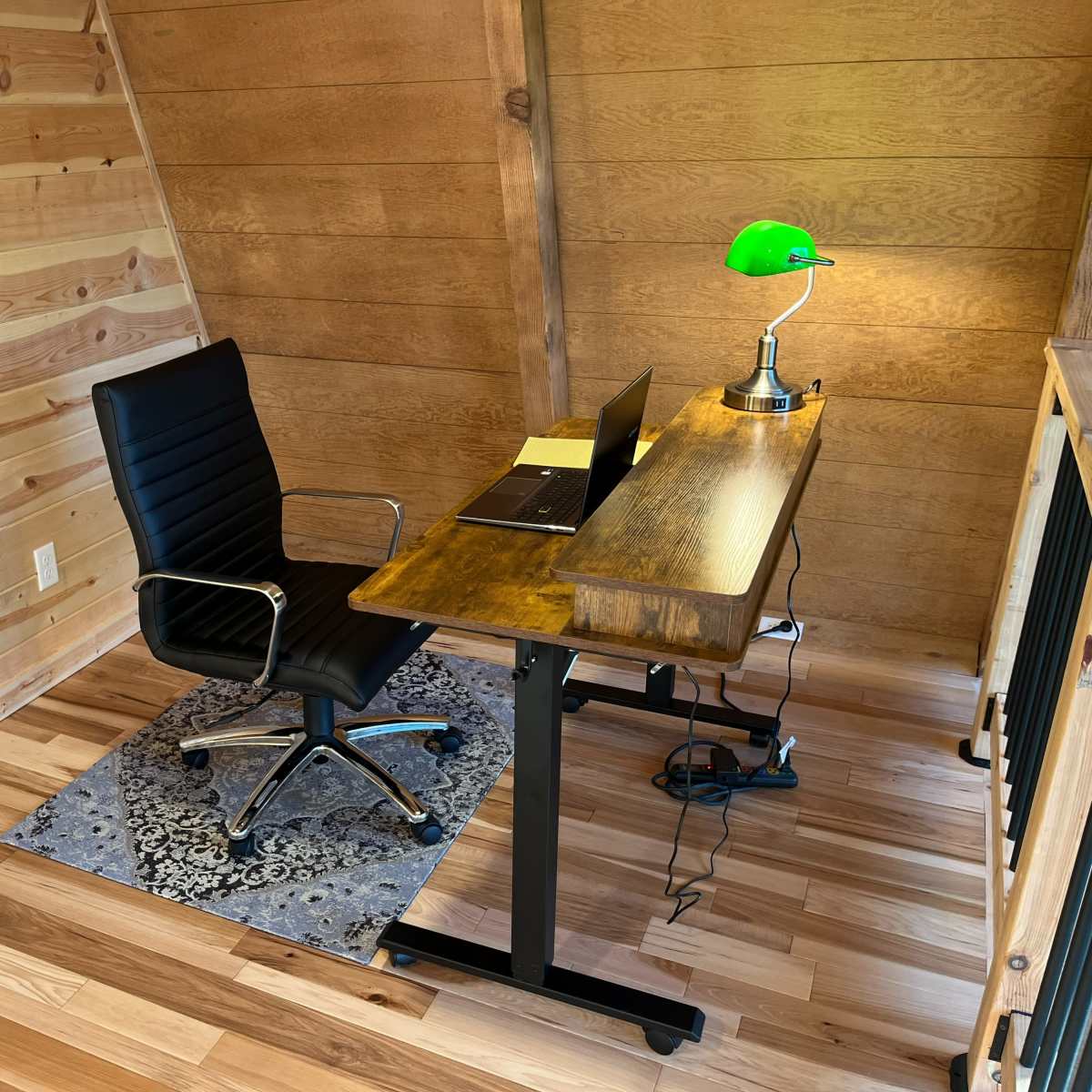 A black ergonomic chair is positioned at a wooden desk with a laptop and a green desk lamp in a room with wooden walls and flooring.