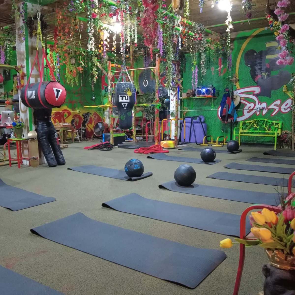 An indoor gym decorated with colorful plants and equipment, featuring exercise mats, punching bags, and fitness balls arranged throughout the space.
