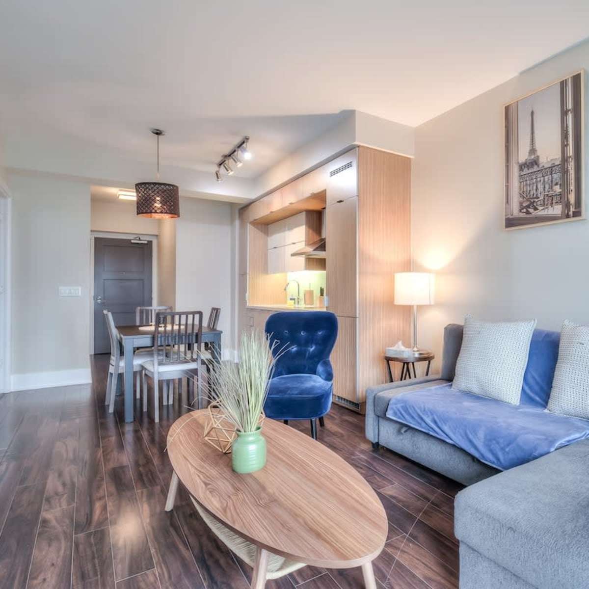 A modern living space featuring a gray sofa, a round wooden coffee table, a dining area with a table and chairs, and wall art in a well-lit room.