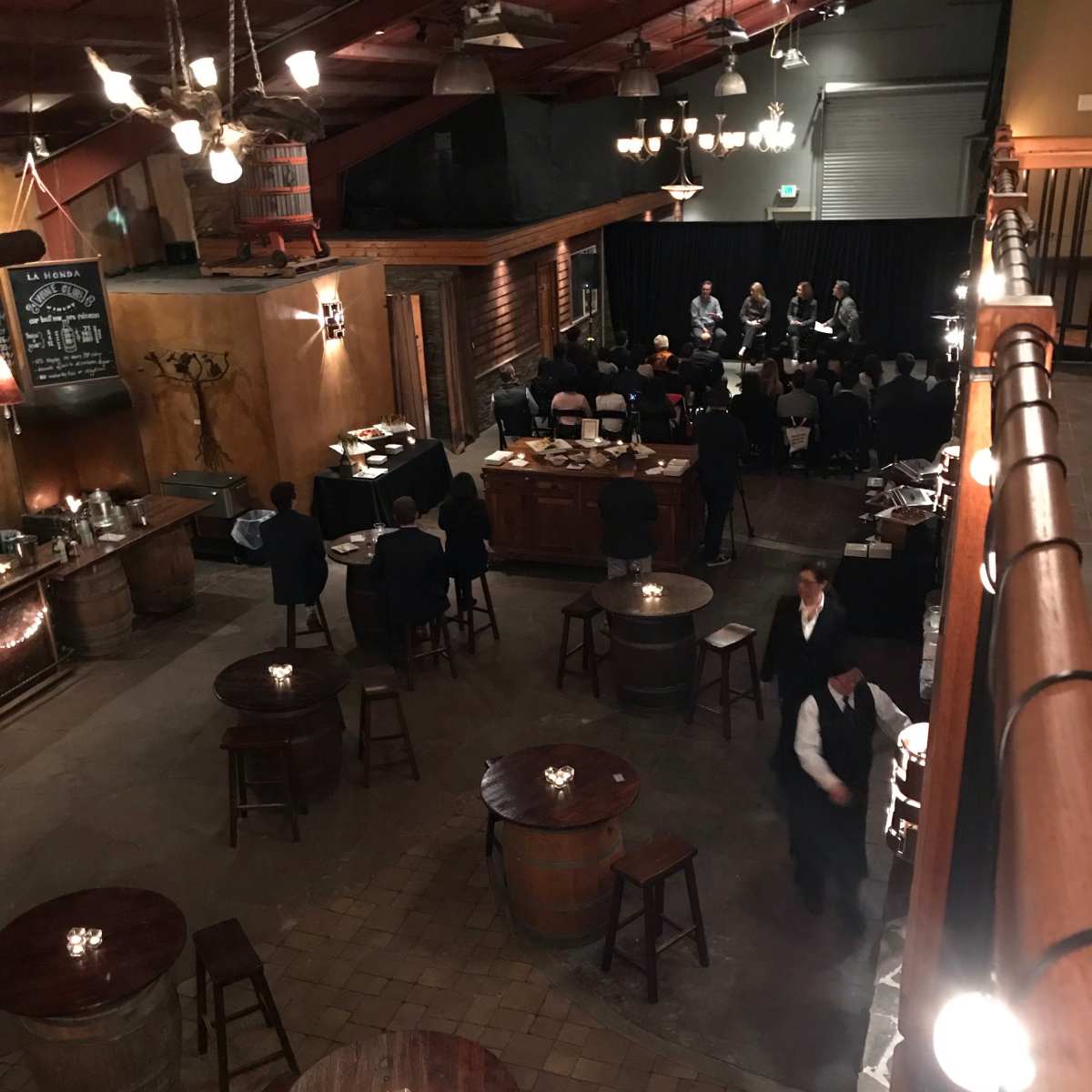 A dimly lit event space with round tables and bar stools, where a group is seated on a stage at the far end.