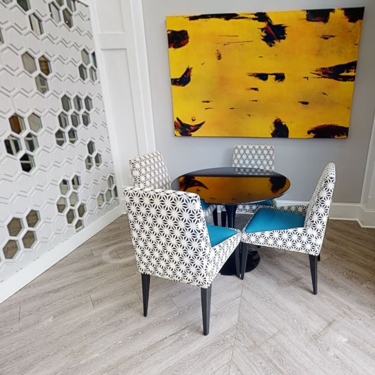 A small dining area features a round black table surrounded by three patterned chairs, with a large yellow abstract painting on the wall.