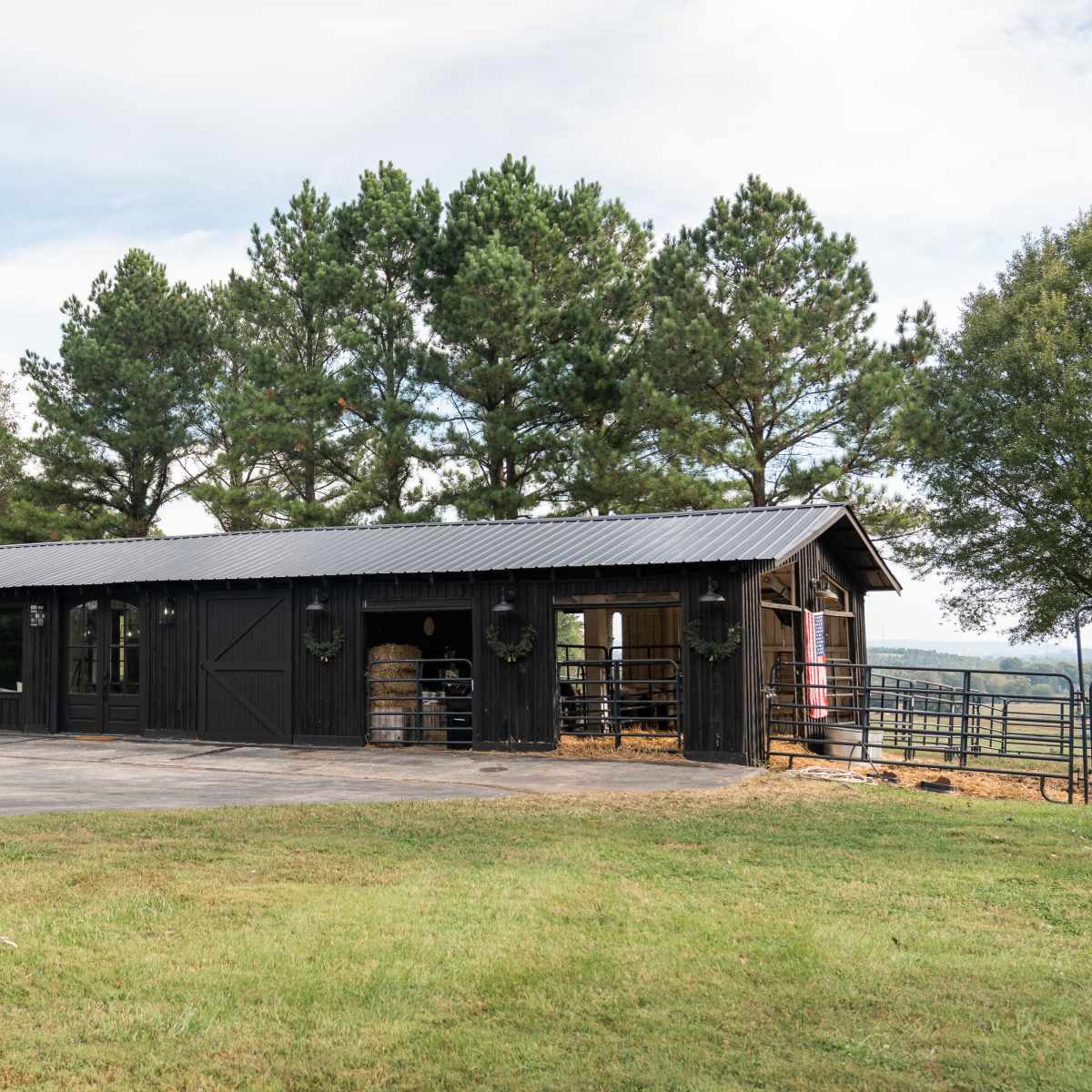 A black barn with large windows and a metal roof sits beside a grassy area, framed by tall trees in the background.
