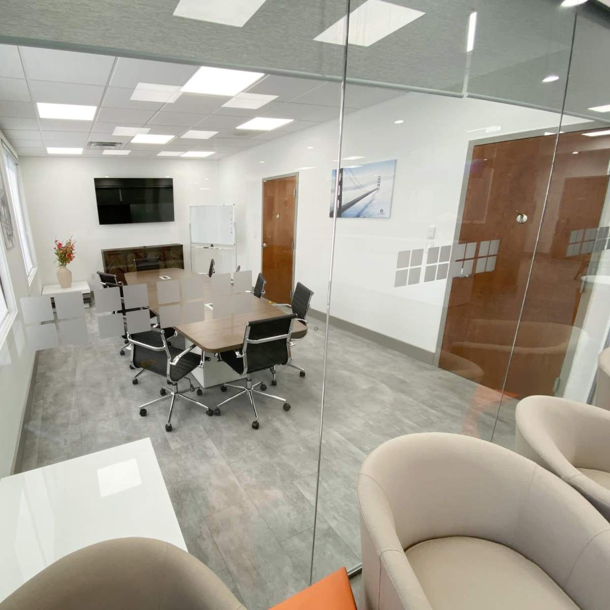 A modern conference room features a large table surrounded by rolling chairs, a TV screen on the wall, and glass partitions.