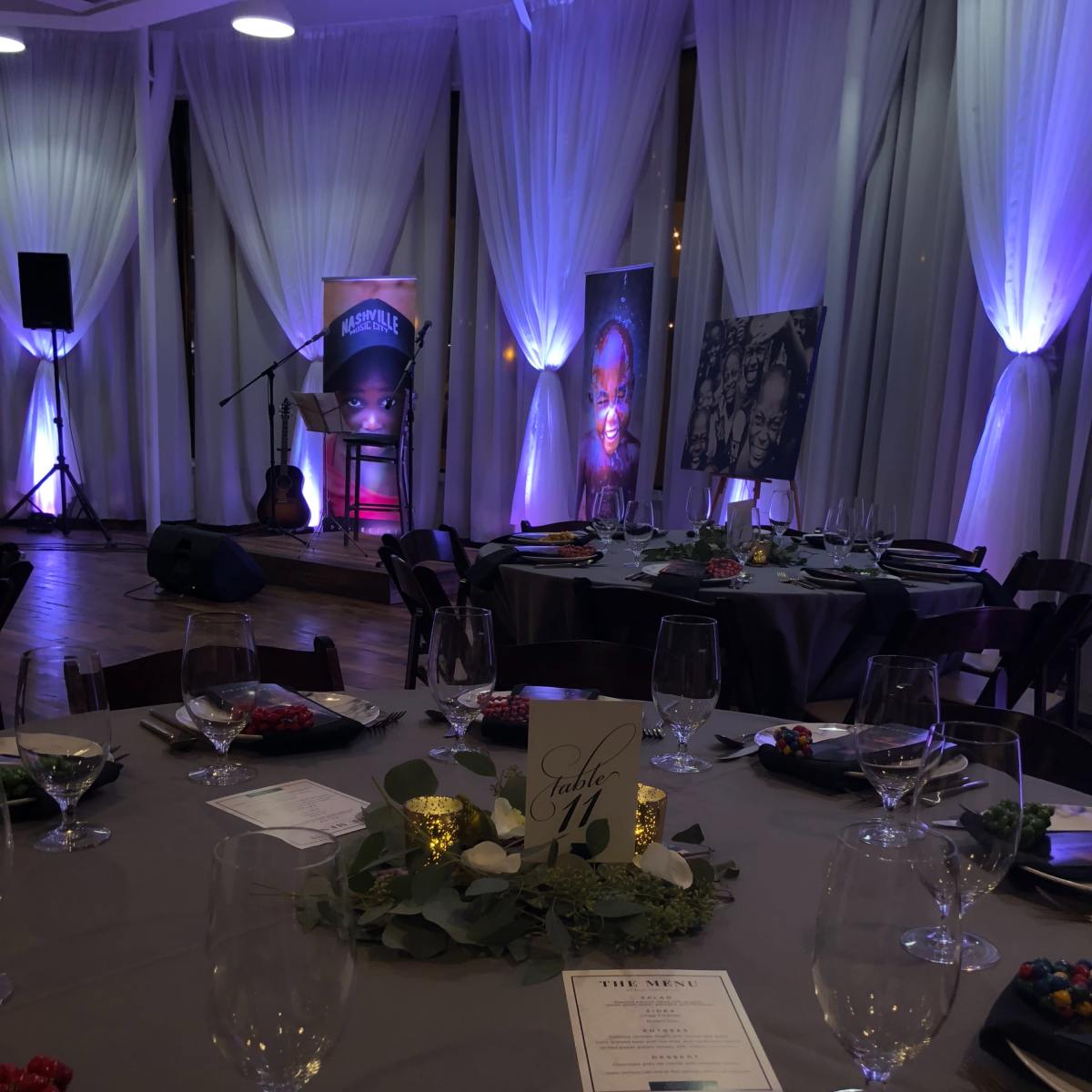 A banquet setup with elegant table settings, including plates, glasses, and a centerpiece, in a room featuring a stage and artwork.