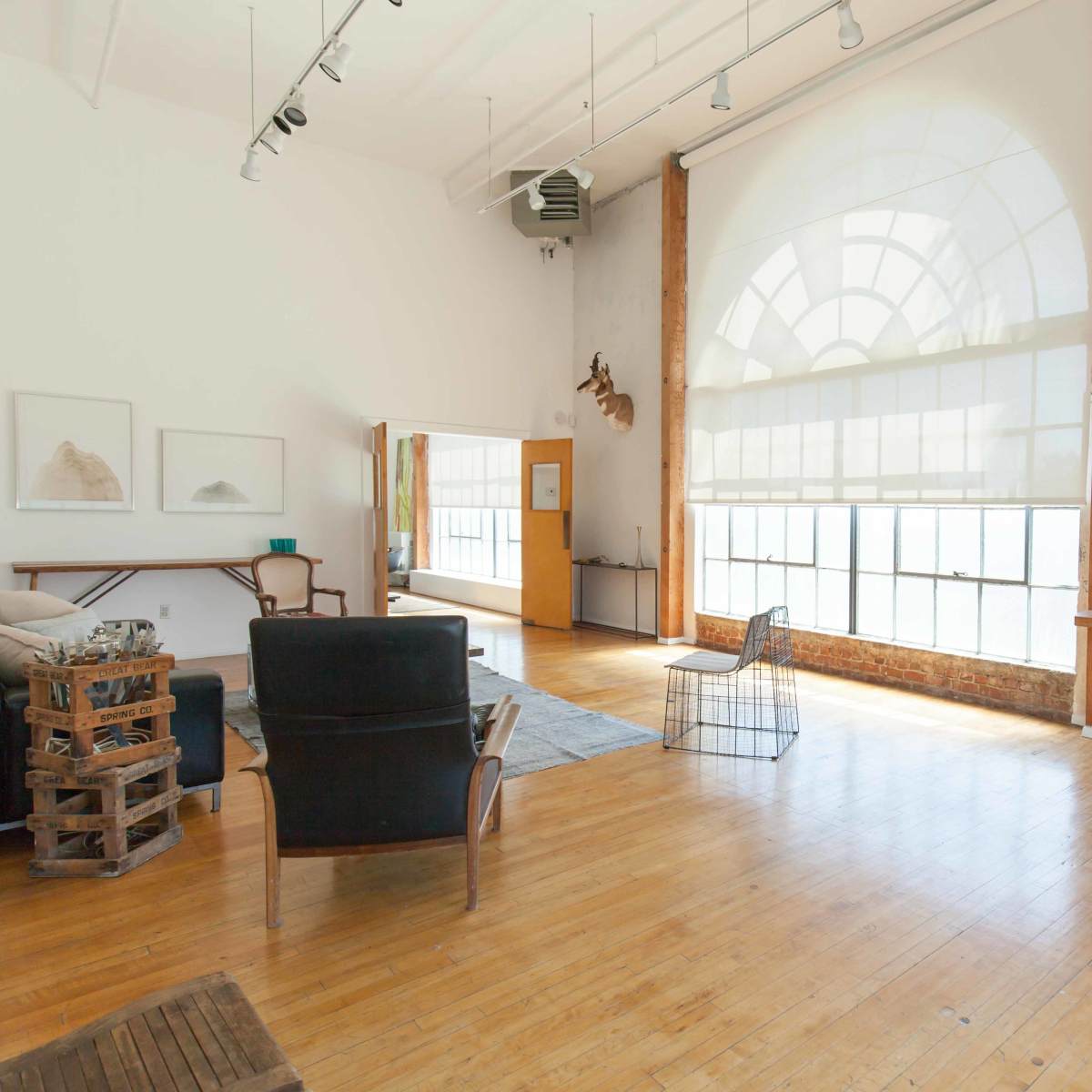 A spacious, brightly lit room with wooden flooring, featuring a large window, bookshelves, and minimalist furniture arrangements.