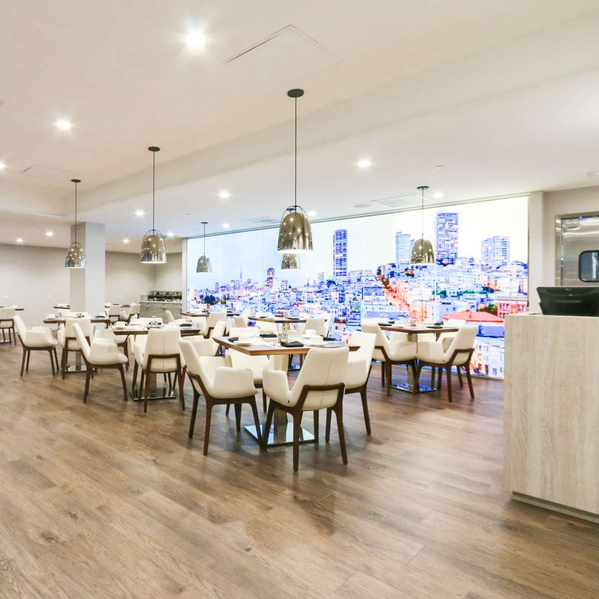 A modern restaurant interior with tables and chairs, illuminated by pendant lights, and featuring a large cityscape mural on one wall.