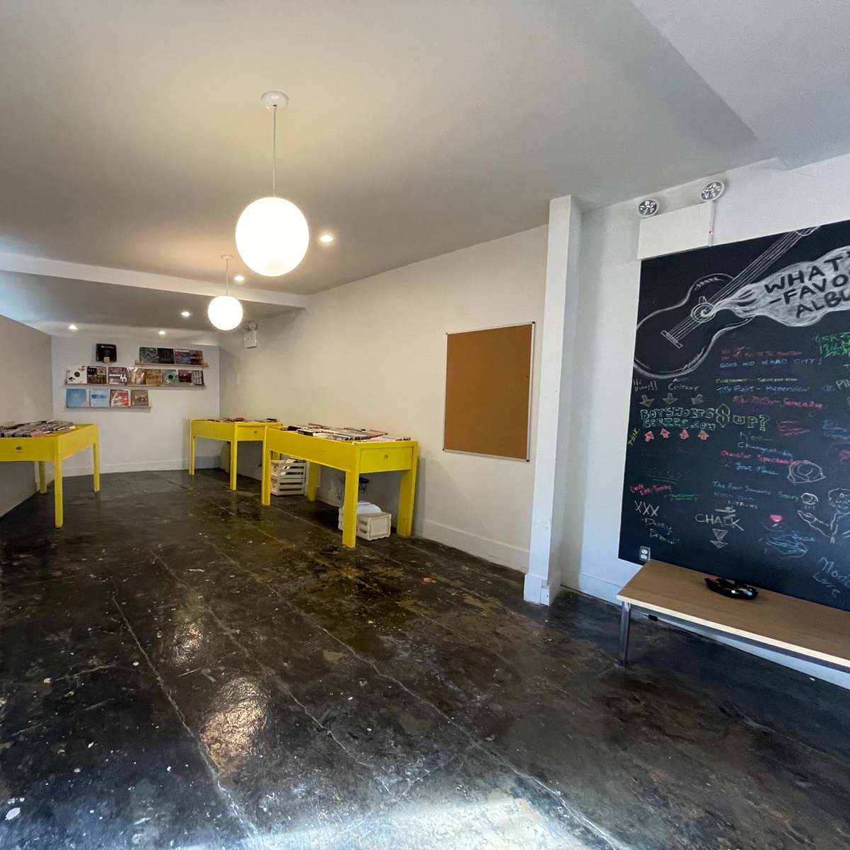 A minimalistic room with yellow tables displaying records, a wall featuring vinyl album covers, and a chalkboard with colorful drawings and writing.