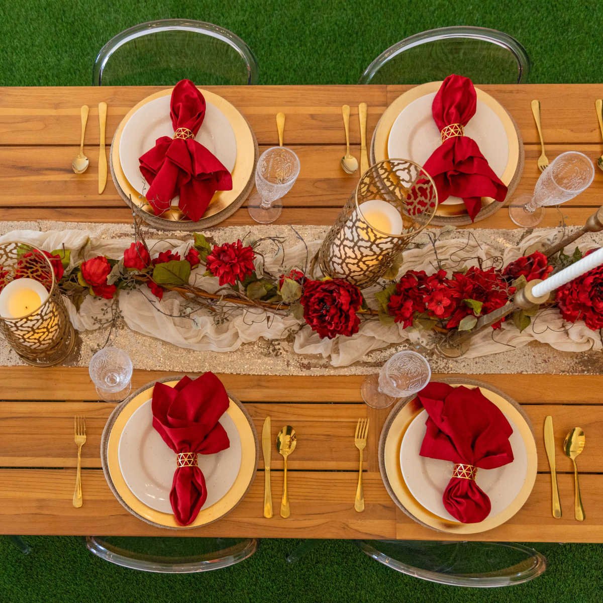 A long wooden dining table is set with gold utensils, white plates, red napkins, and decorative centerpieces featuring candles and flowers.