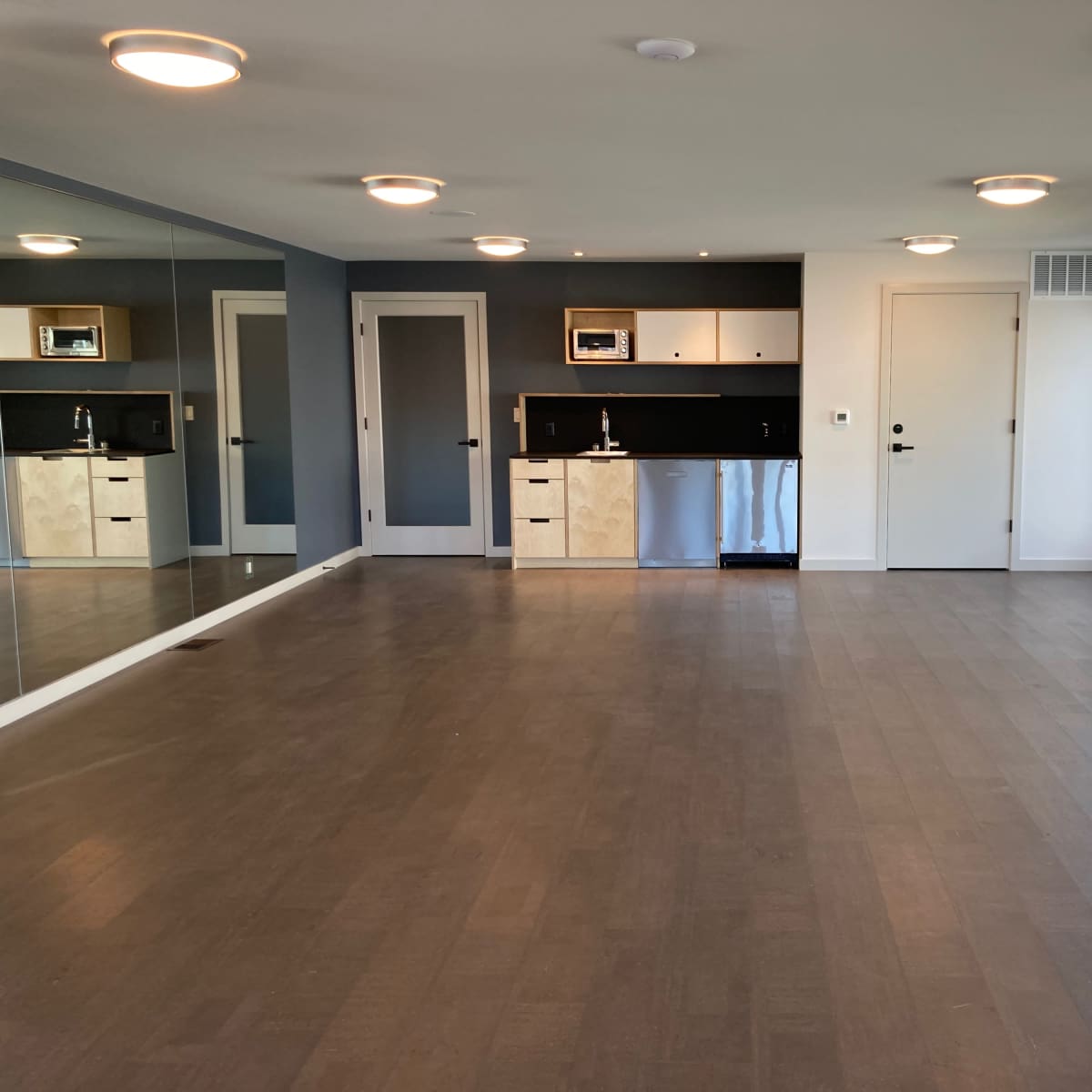 A spacious, modern room with a mirrored wall, light fixtures on the ceiling, a small kitchenette in the corner, and large windows providing natural light.