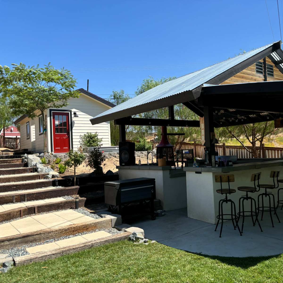 A covered outdoor bar with several stools, adjacent to a house and landscaped steps leading to it.