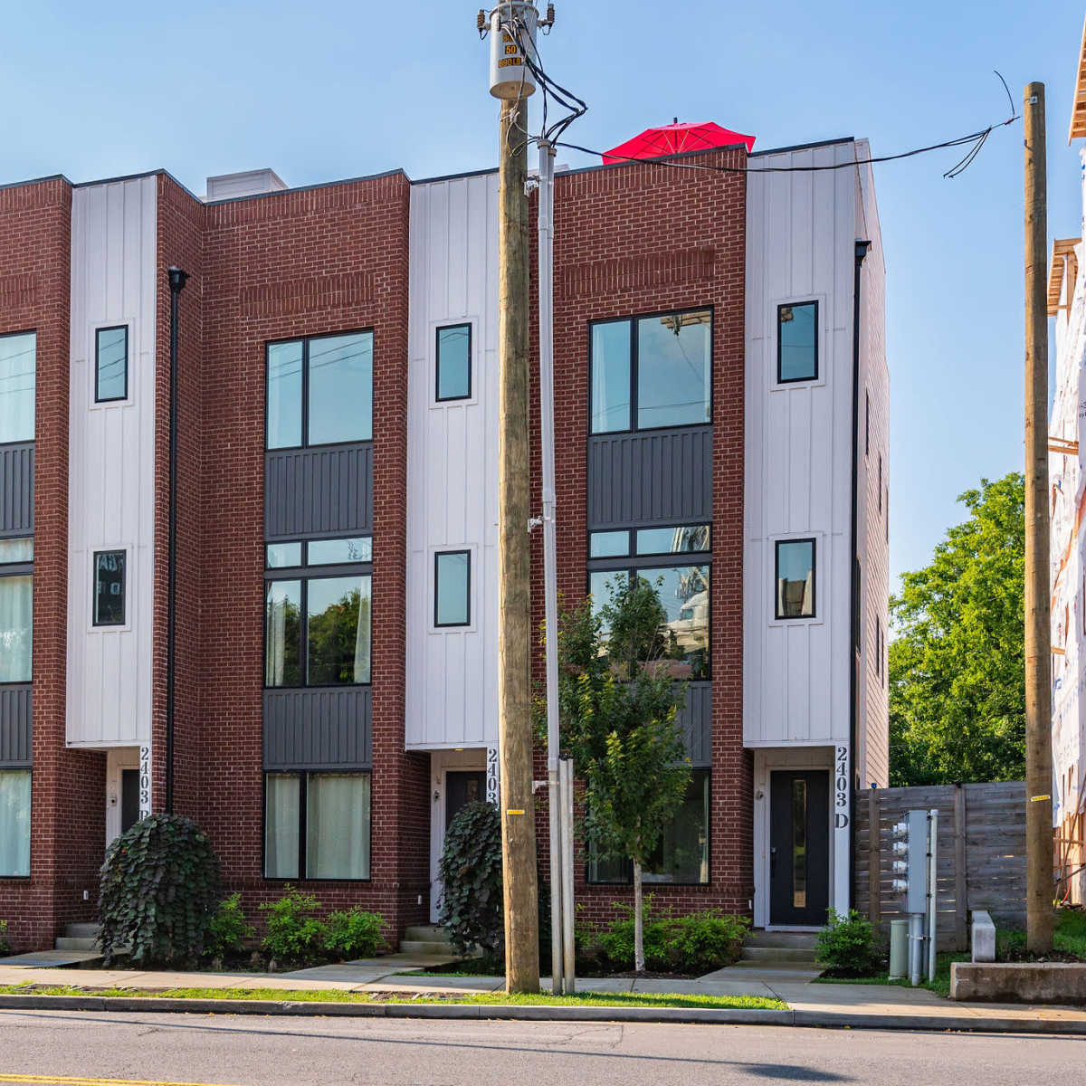 A modern, three-story brick building with large windows and a red umbrella on the roof, situated on a street with decorative landscaping.