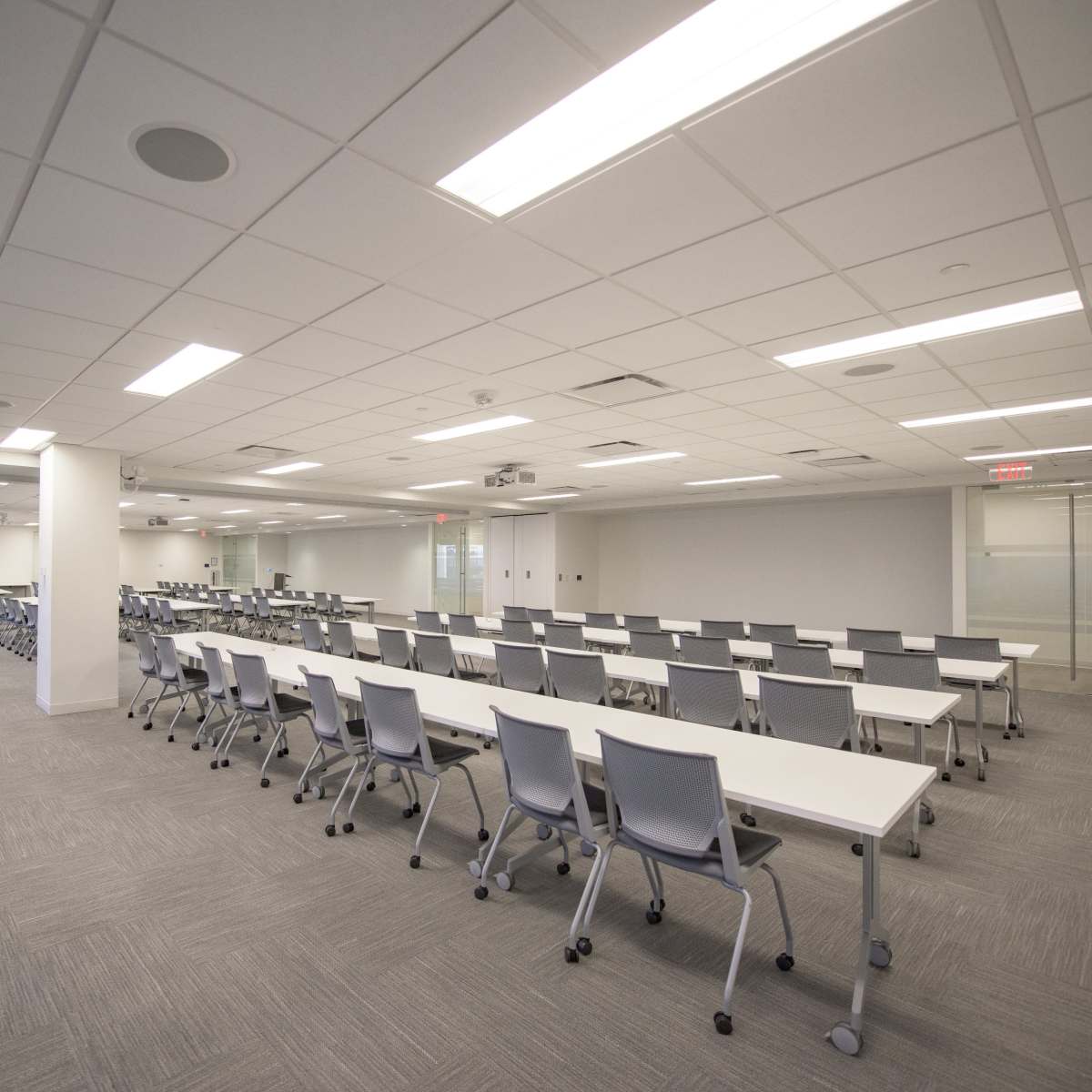 A spacious, modern conference room with multiple rows of white tables and gray chairs arranged neatly.