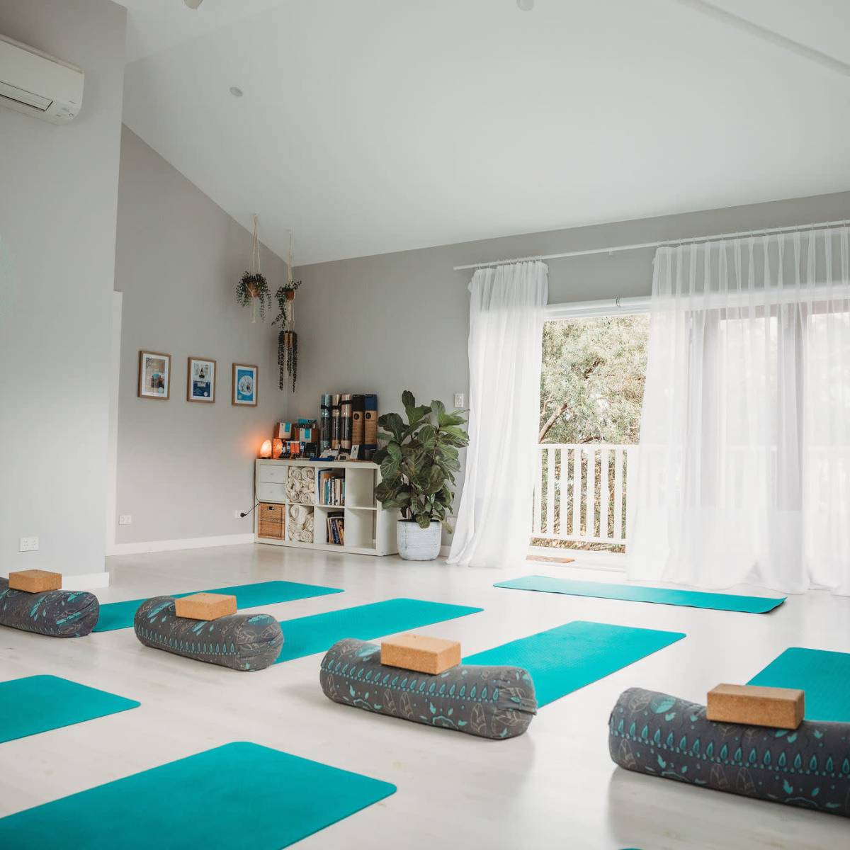 A spacious yoga studio is set up with green mats and bolsters facing a large window, with plants and decor lining the walls.