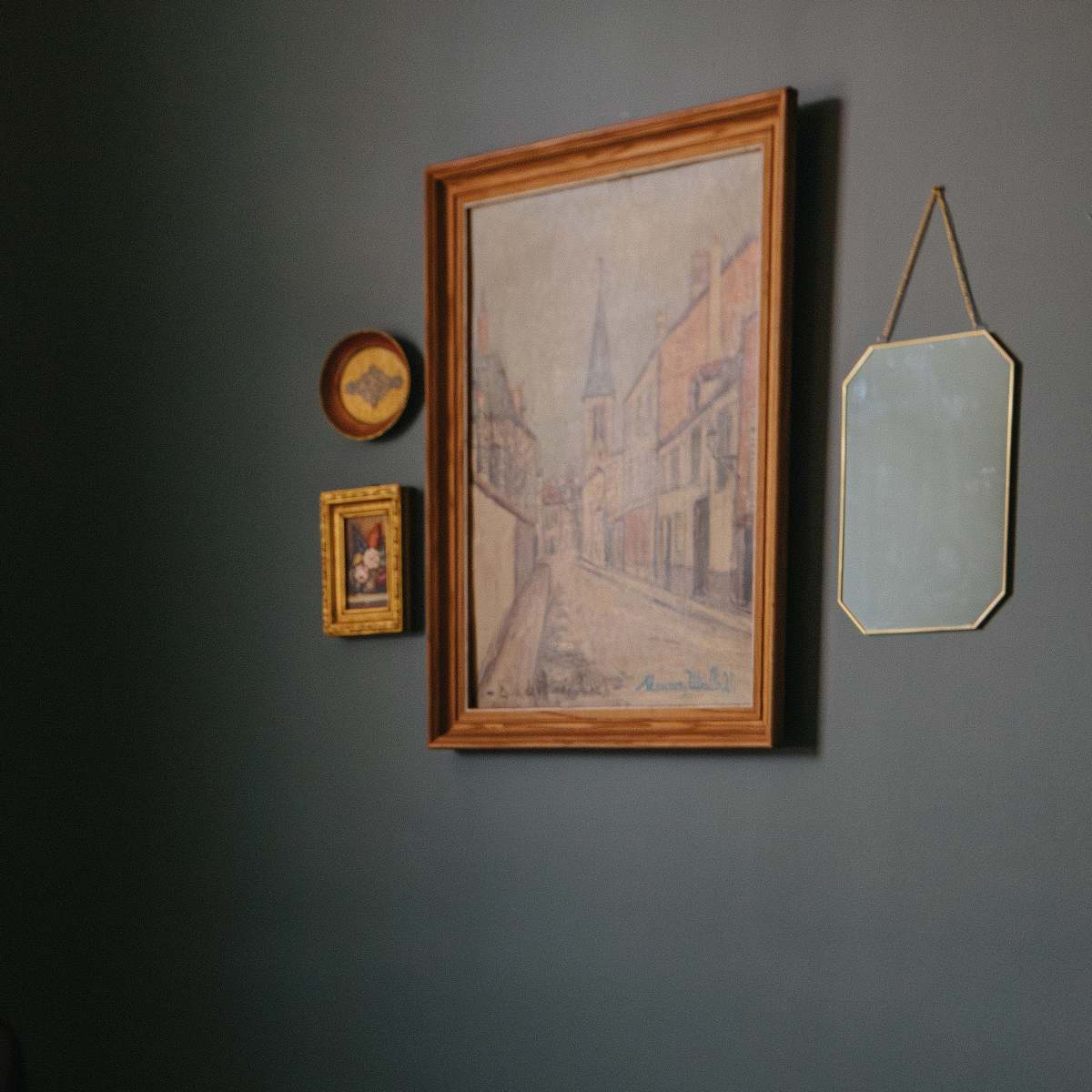 A wall painted in a dark color adorned with a large painting, two smaller framed artworks, and an octagonal mirror.