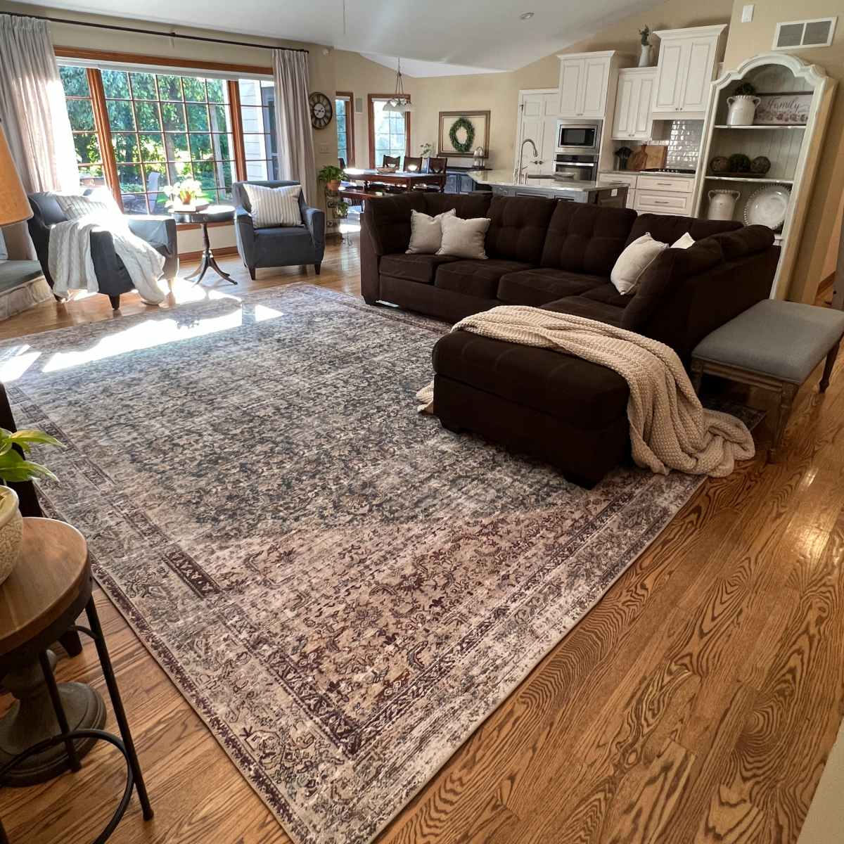 A spacious living room with a large dark sectional couch, a decorative rug, and a bright window overlooking a garden.