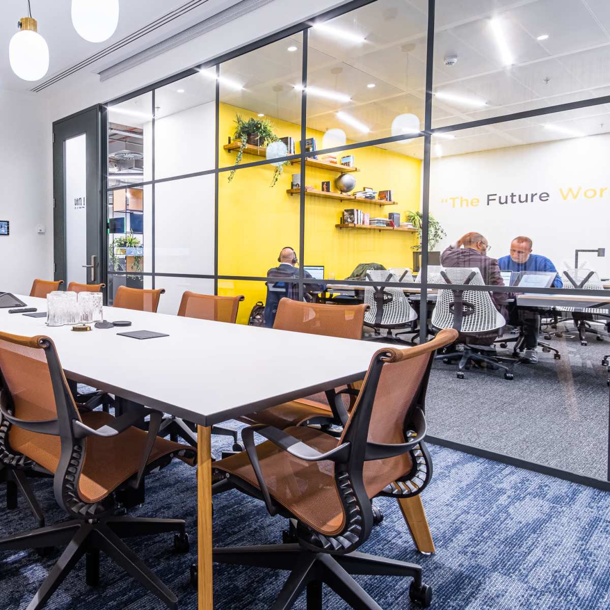 A modern conference room features a long table surrounded by ergonomic chairs, with a screen displaying a cityscape on one wall and colleagues discussing behind a glass partition.