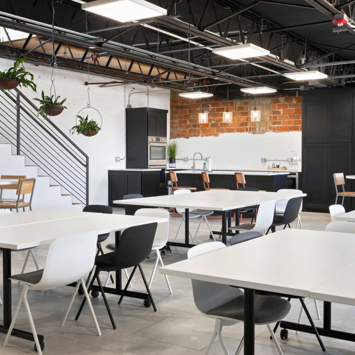 A modern office kitchen and dining area with white tables and multi-colored chairs under exposed ceiling beams and brick walls.