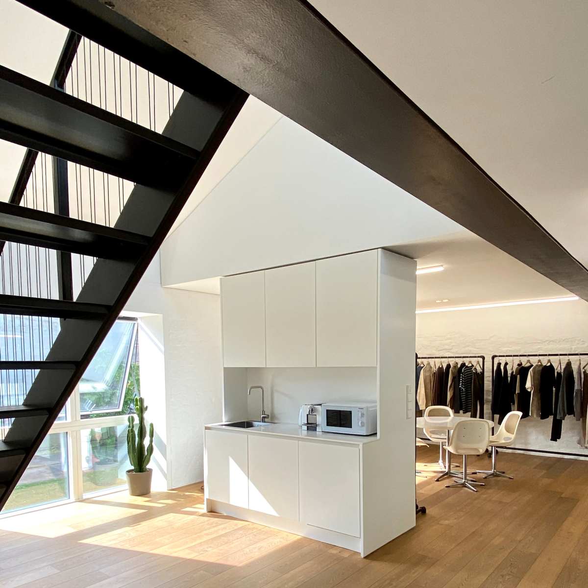 A modern kitchen with white cabinetry and a microwave, alongside a clothing rack with various garments in a well-lit room featuring wooden flooring.