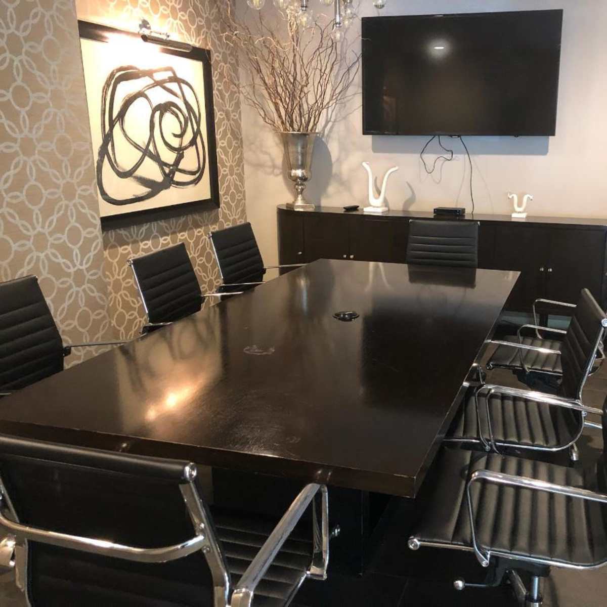A modern conference room with a large rectangular table surrounded by black chairs, featuring a wall-mounted TV and decorative wall art.