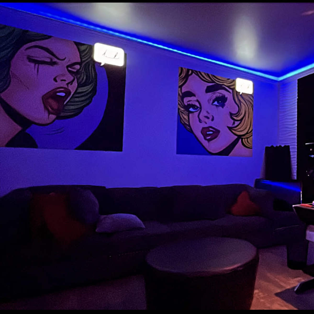 A modern music studio with colorful wall art, a dark couch, and a desk equipped with audio equipment under blue LED lighting.