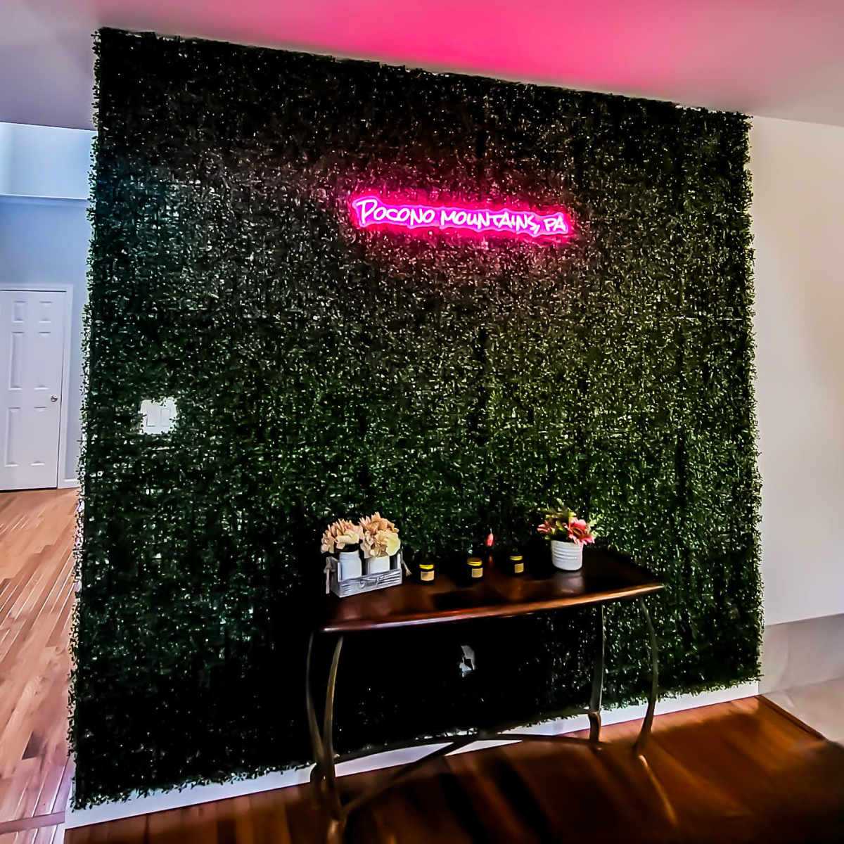 A wall covered in greenery with a neon sign reading "Pocono Mountains PA" and a wooden table displaying decorative items in front of it.
