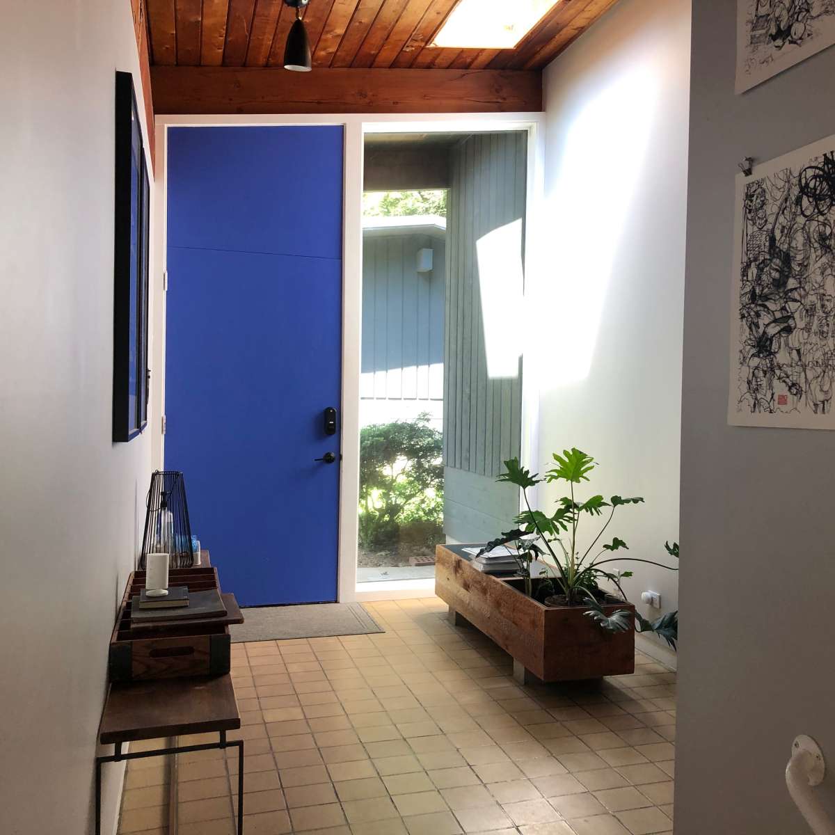 A hallway with a blue front door, a wooden planter with plants, and artwork hanging on the walls.