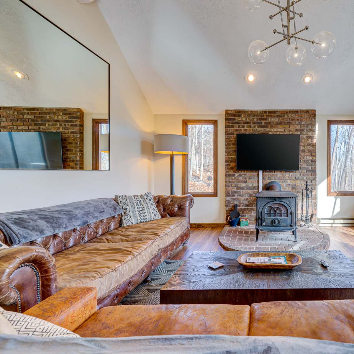 A modern living room featuring a leather sofa, a wooden coffee table, a wood-burning stove, and large windows overlooking a wooded area.
