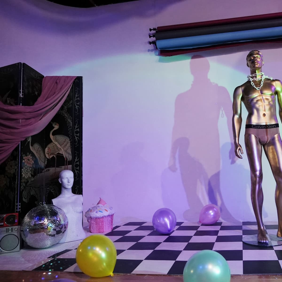 The scene features a metallic mannequin in underwear standing next to a microphone on a checkered floor, surrounded by colorful balloons and a decorative backdrop.