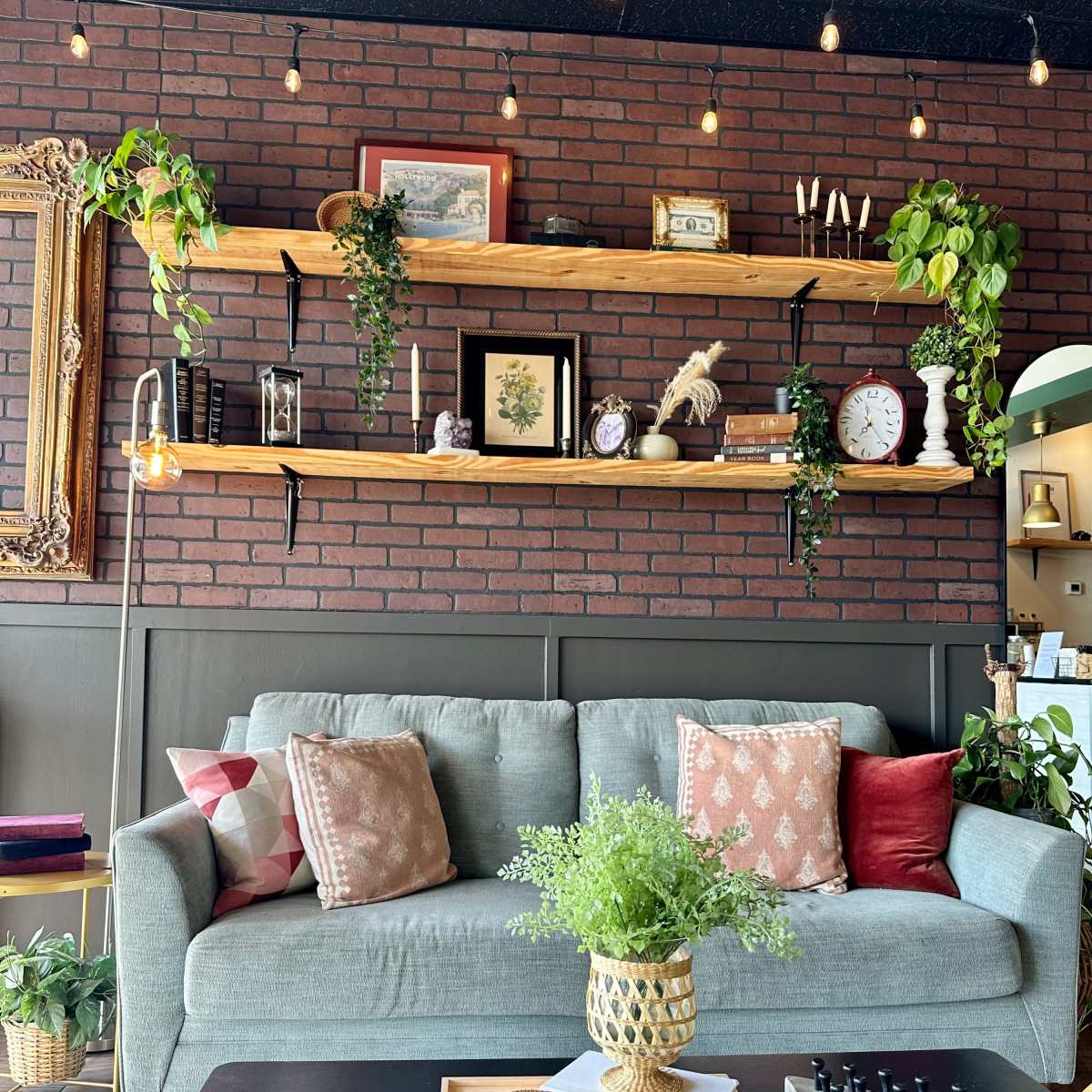 A cozy living area features a green sofa adorned with decorative pillows, placed beneath wooden shelves lined with plants and framed pictures against a brick wall.